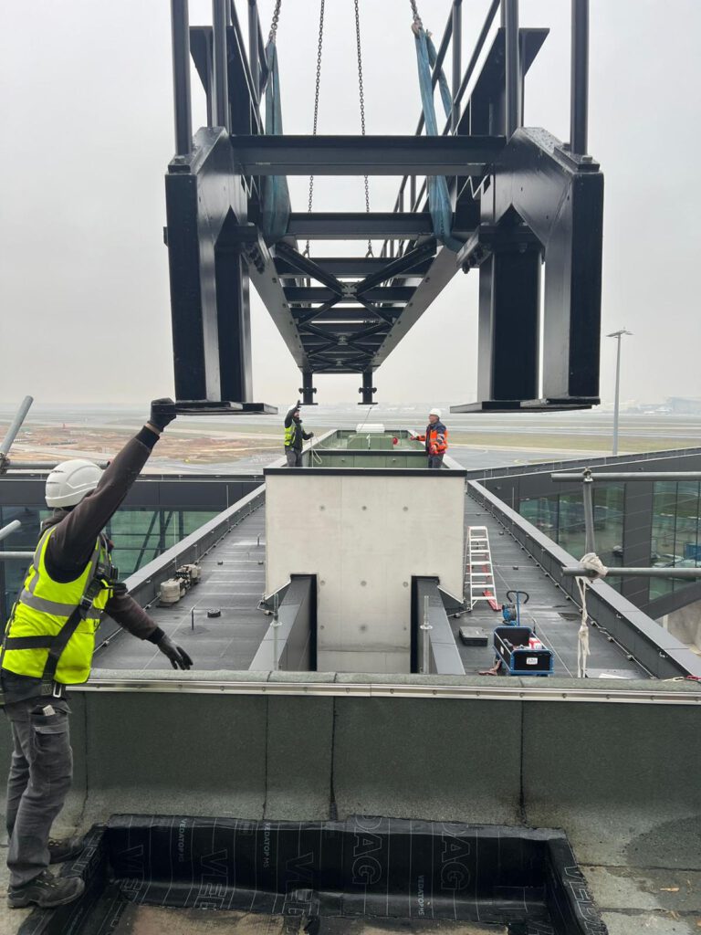 Eine große Metallkonstruktion wird von einem Kran auf das Dach eines Gebäudes gehoben, während drei Arbeiter in Schutzausrüstung die Installation an einem bewölkten Tag leiten.