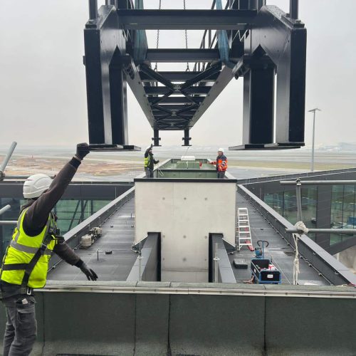Eine große Metallkonstruktion wird von einem Kran auf das Dach eines Gebäudes gehoben, während drei Arbeiter in Schutzausrüstung die Installation an einem bewölkten Tag leiten.