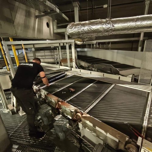 Eine Person in Arbeitskleidung inspiziert oder repariert ein großes Fördersystem in einer Industrieanlage, in der verschiedene Rohre, Metallstrukturen und Maschinen sichtbar sind.